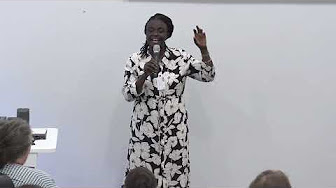 Professor Olaniyi standing, wearing a black and white dress and holding a microphone